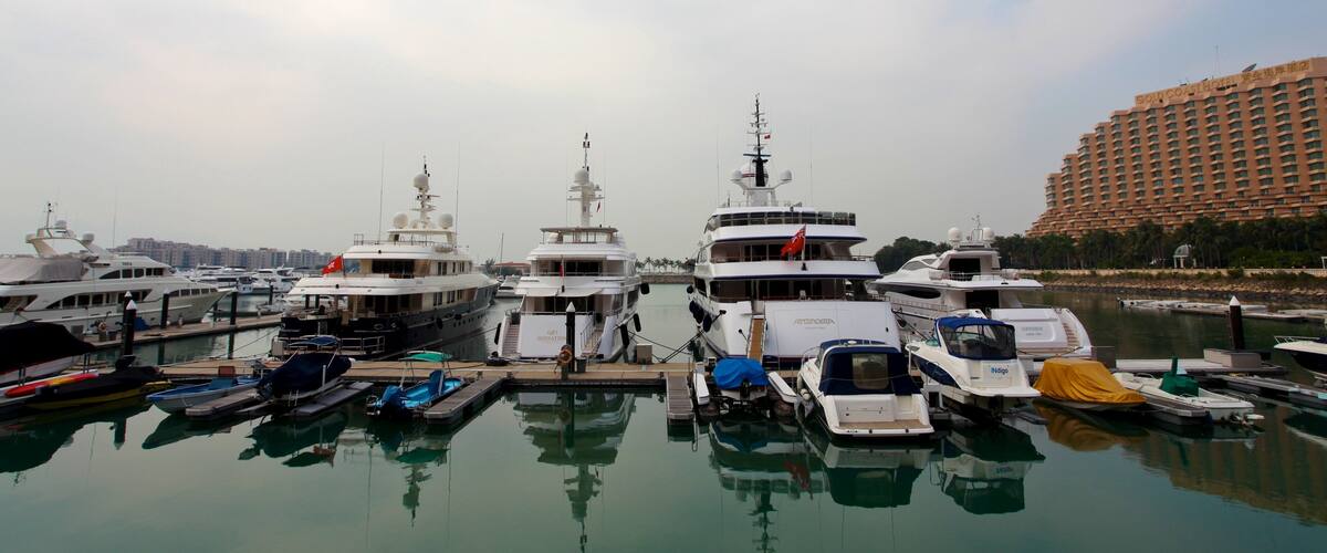 A marina with multiple yachts docked, a large hotel in the background, and calm water reflecting the boats.