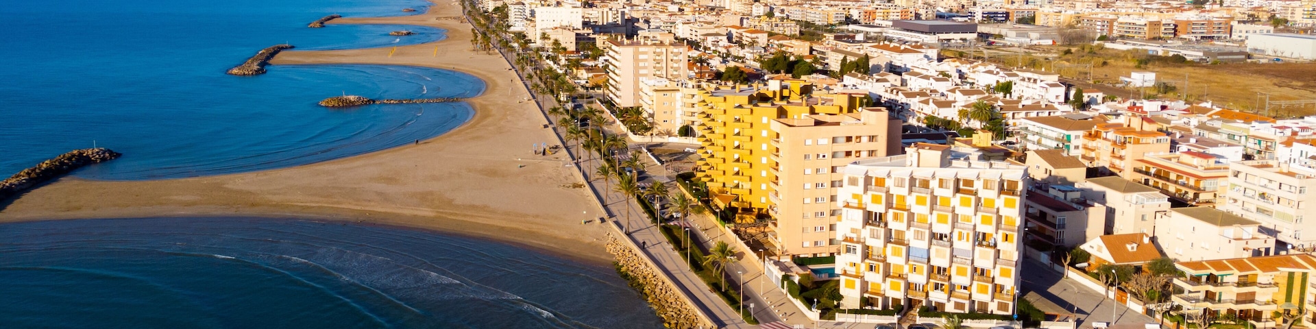 A coastal cityscape with a wide beach, a rocky breakwater, and densely packed buildings along the shore.