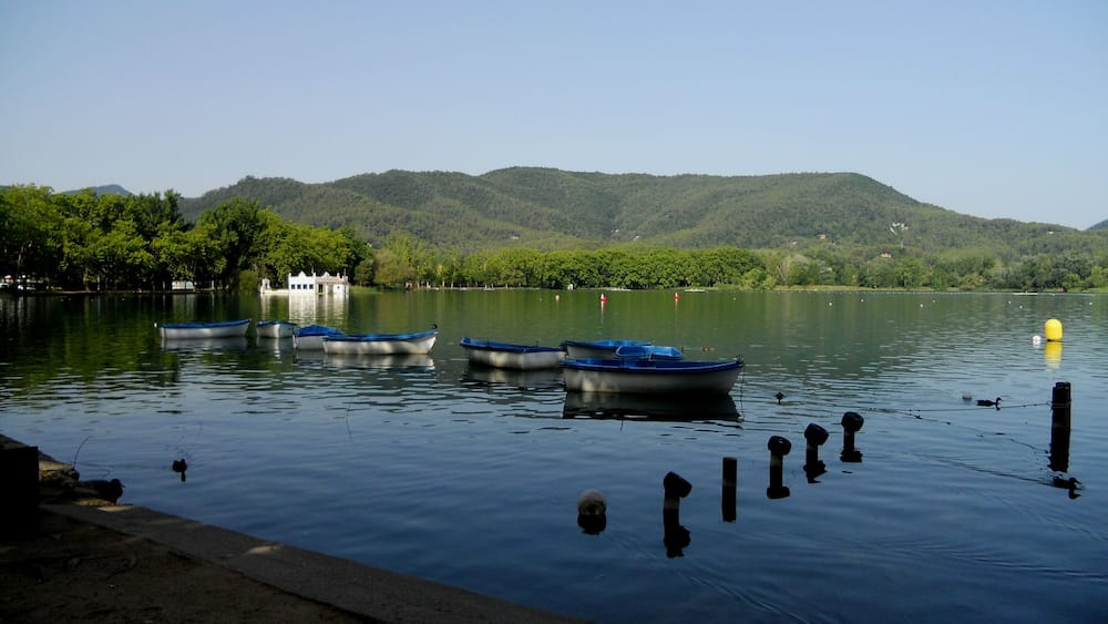 A calm lake with several small boats docked near the shore, surrounded by lush green hills and a clear sky.