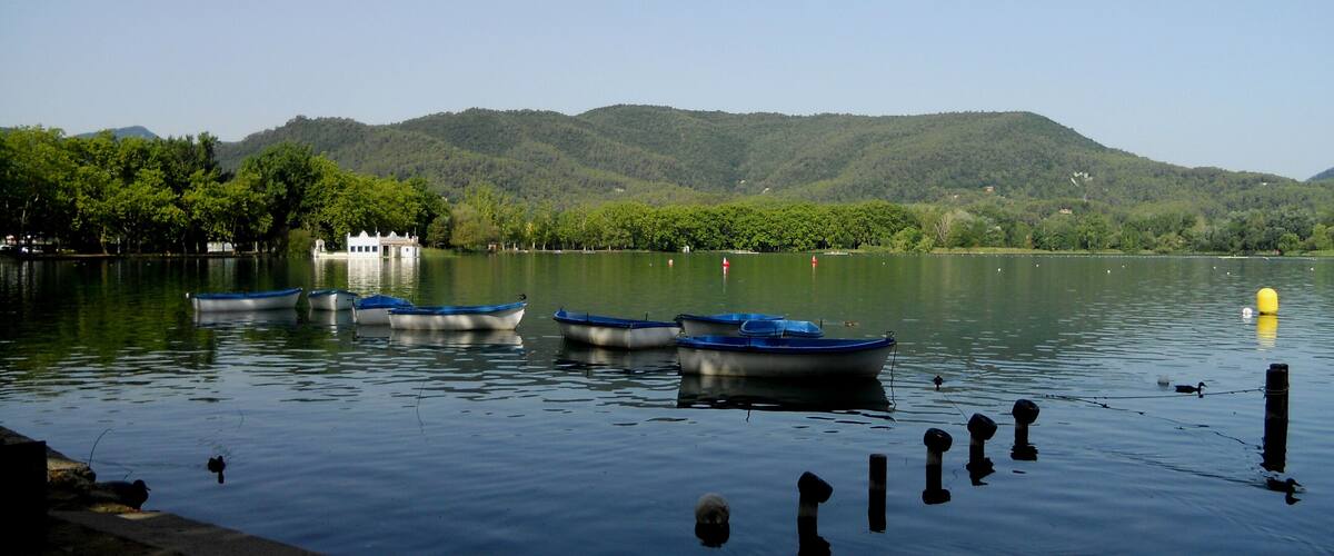 A calm lake with several small boats docked near the shore, surrounded by lush green hills and a clear sky.
