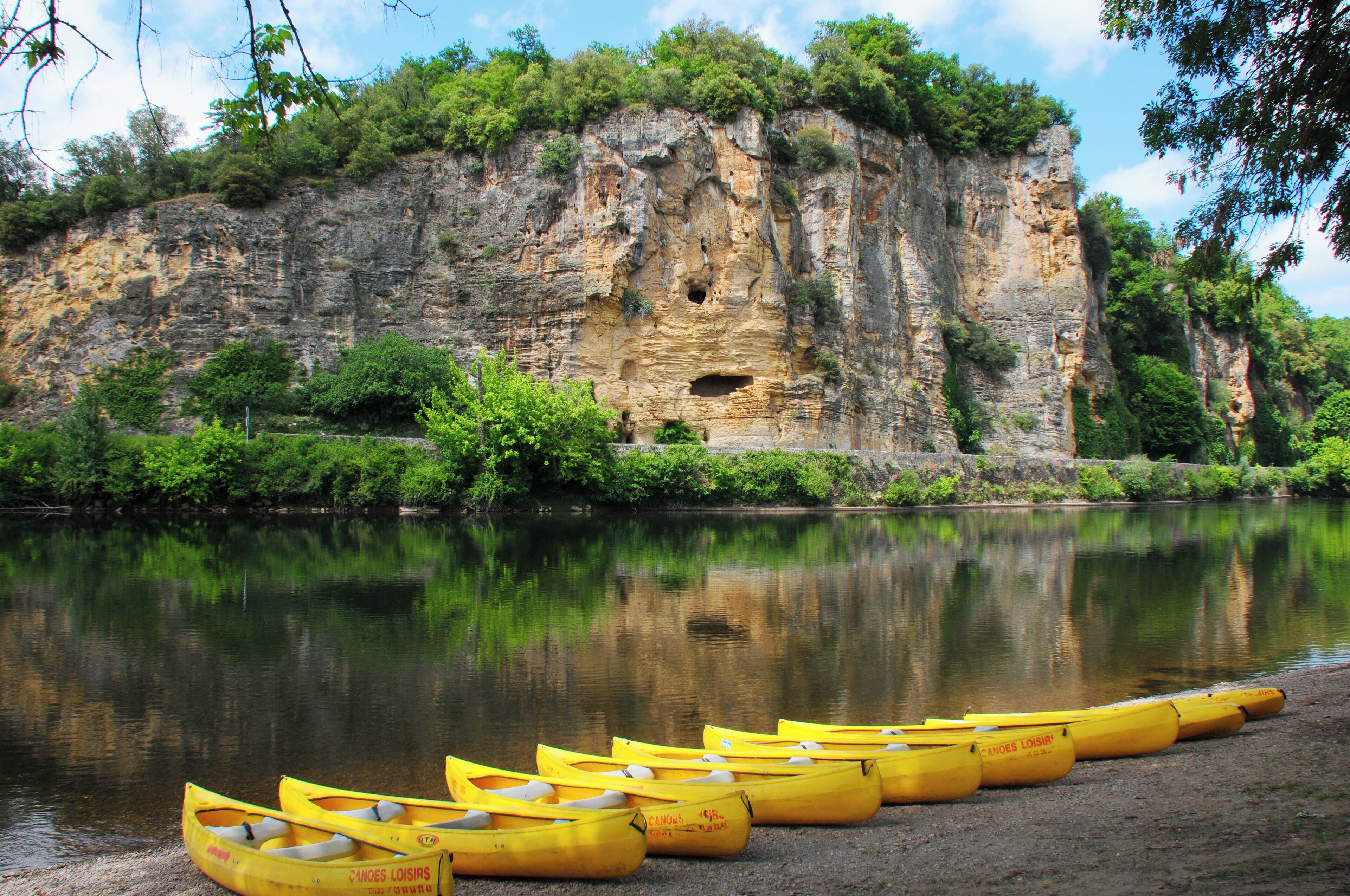 A river with yellow canoes lined up on the shore, a rocky cliff with a cave, and lush greenery.