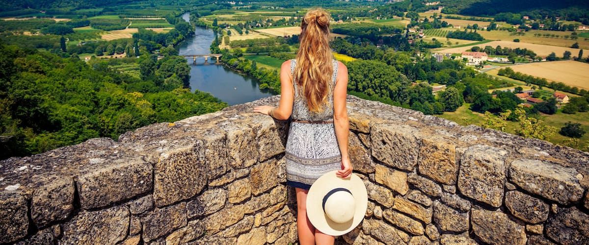 A woman overlooking a river valley with a stone wall, greenery, and distant buildings.