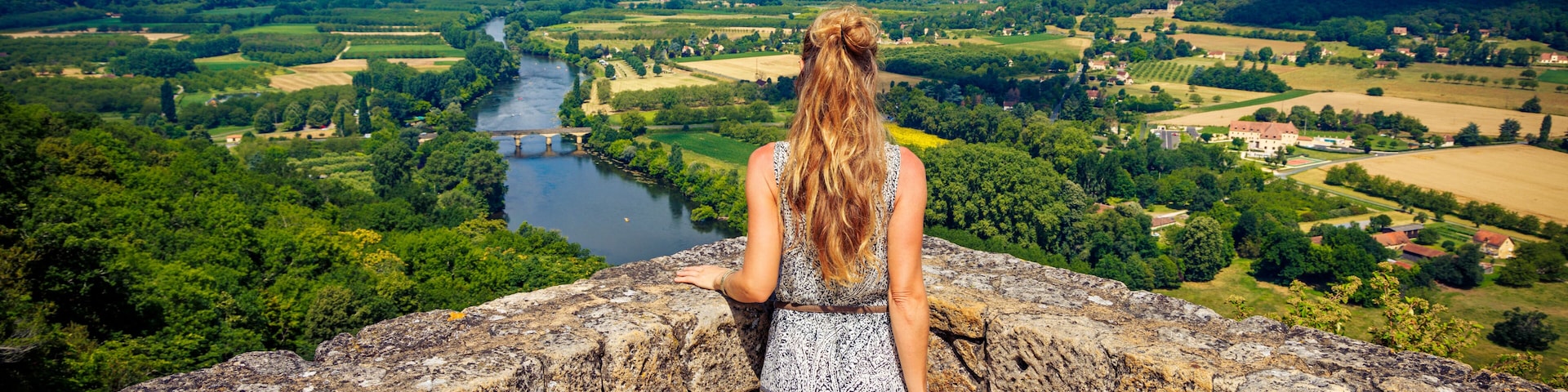 A woman overlooking a river valley with a stone wall, greenery, and distant buildings.