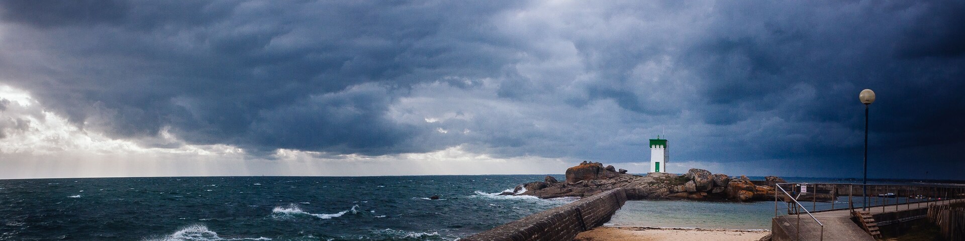 A rocky pier with a lighthouse, turbulent sea, and stormy sky.