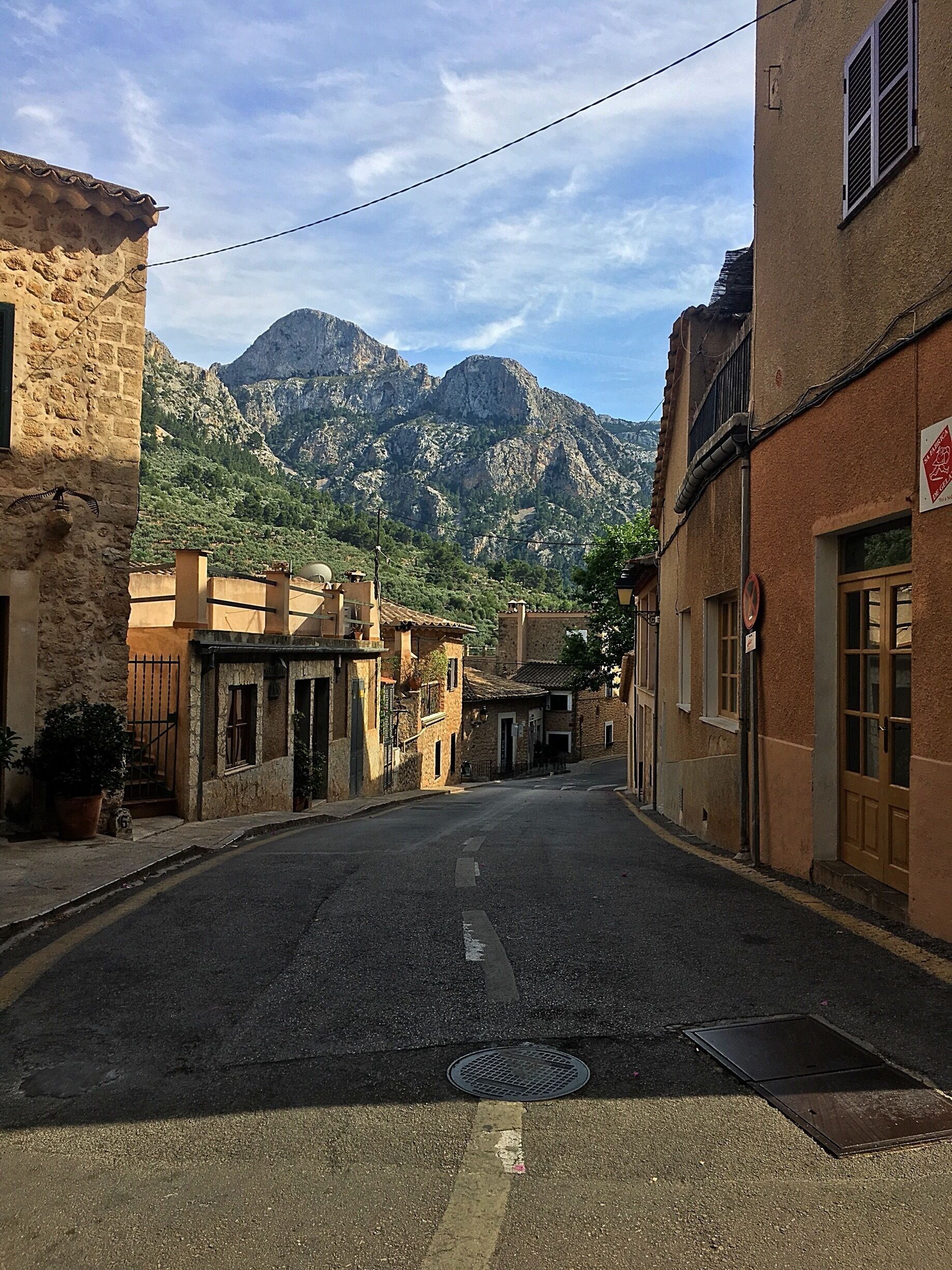 A narrow street flanked by stone buildings, a mountain range in the background, and a clear sky.