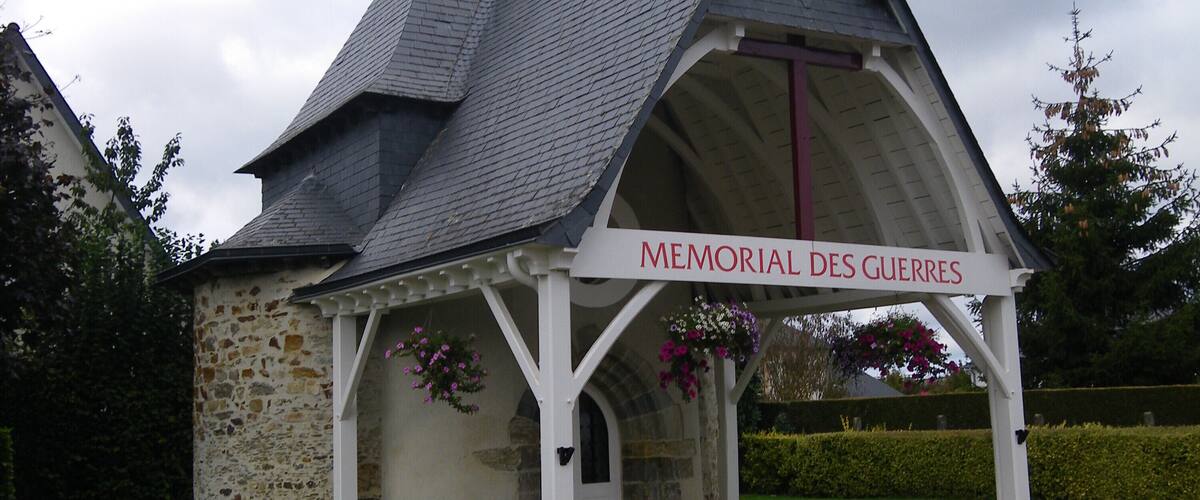 A small stone chapel with a white roof and a sign reading "Memorial Des Guerres".