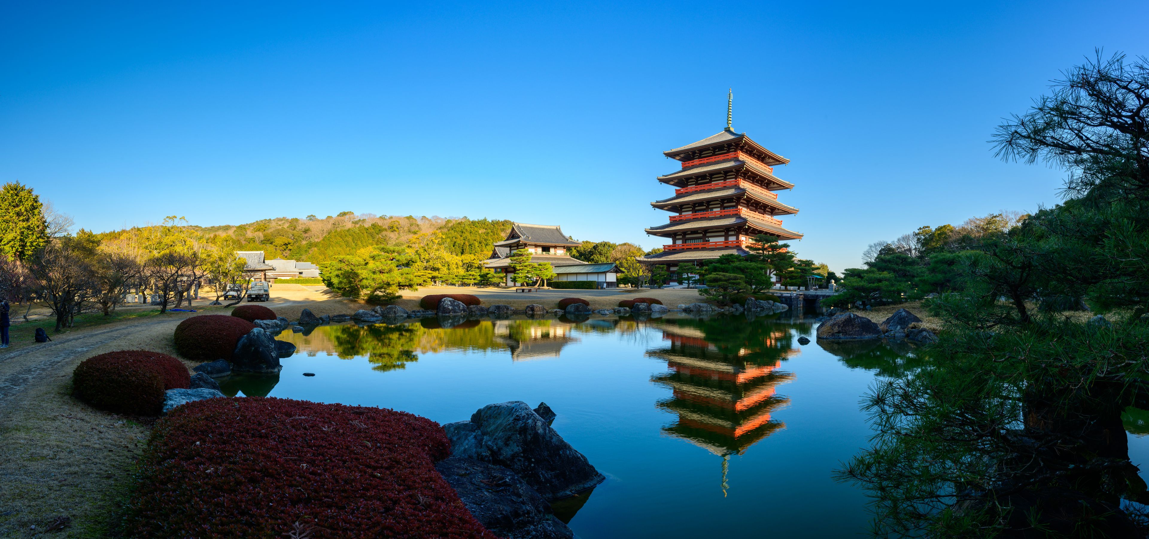 Ett traditionellt japanskt pagodtorn som speglas i vattnet, omgivet av vackert anlagda trädgårdar och en klarblå himmel.