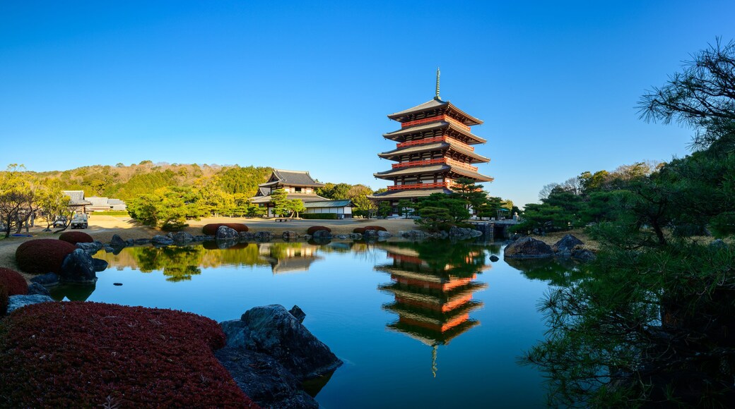 Ett traditionellt japanskt pagodtorn som speglas i vattnet, omgivet av vackert anlagda trädgårdar och en klarblå himmel.