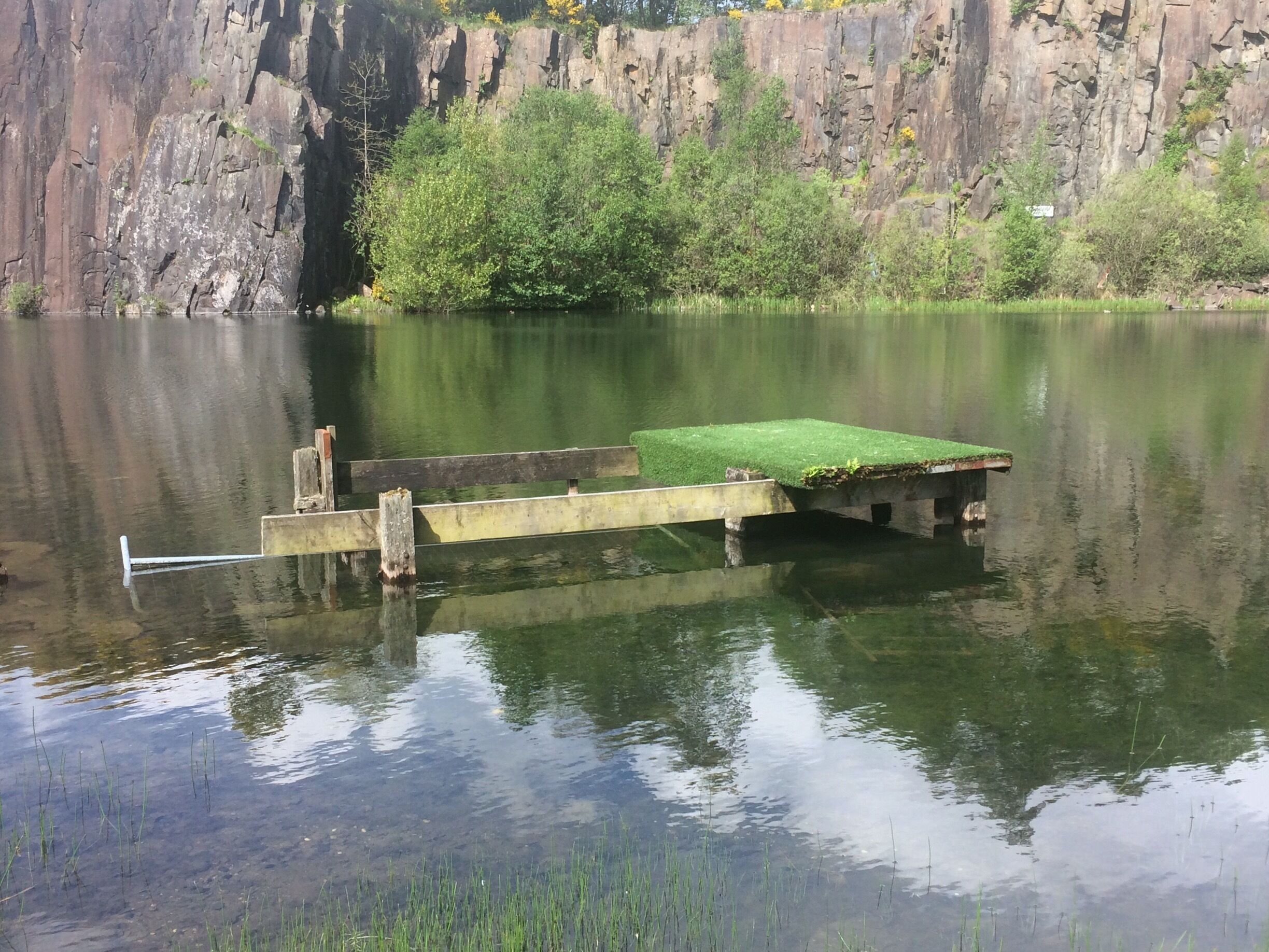 A calm lake with a wooden dock extending into the water, surrounded by rocky cliffs and greenery.