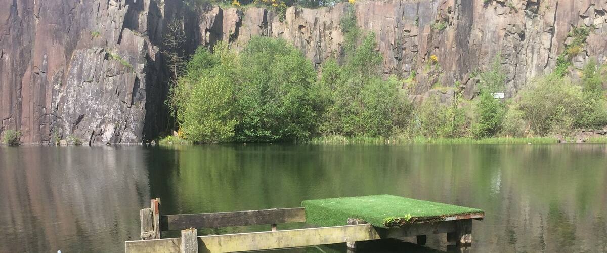 A calm lake with a wooden dock extending into the water, surrounded by rocky cliffs and greenery.