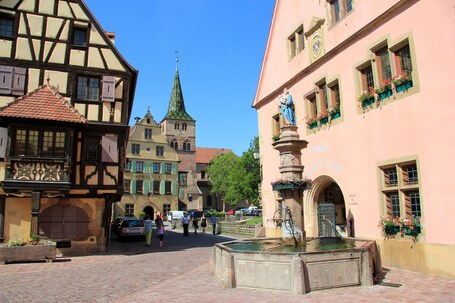 Ein historischer europäischer Stadtplatz mit Fachwerkhäusern, einem Kirchturm und einem Brunnen.