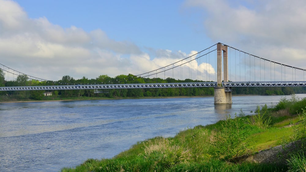 Un pont suspendu enjambant une rivière, avec une végétation luxuriante et des arbres en arrière-plan.