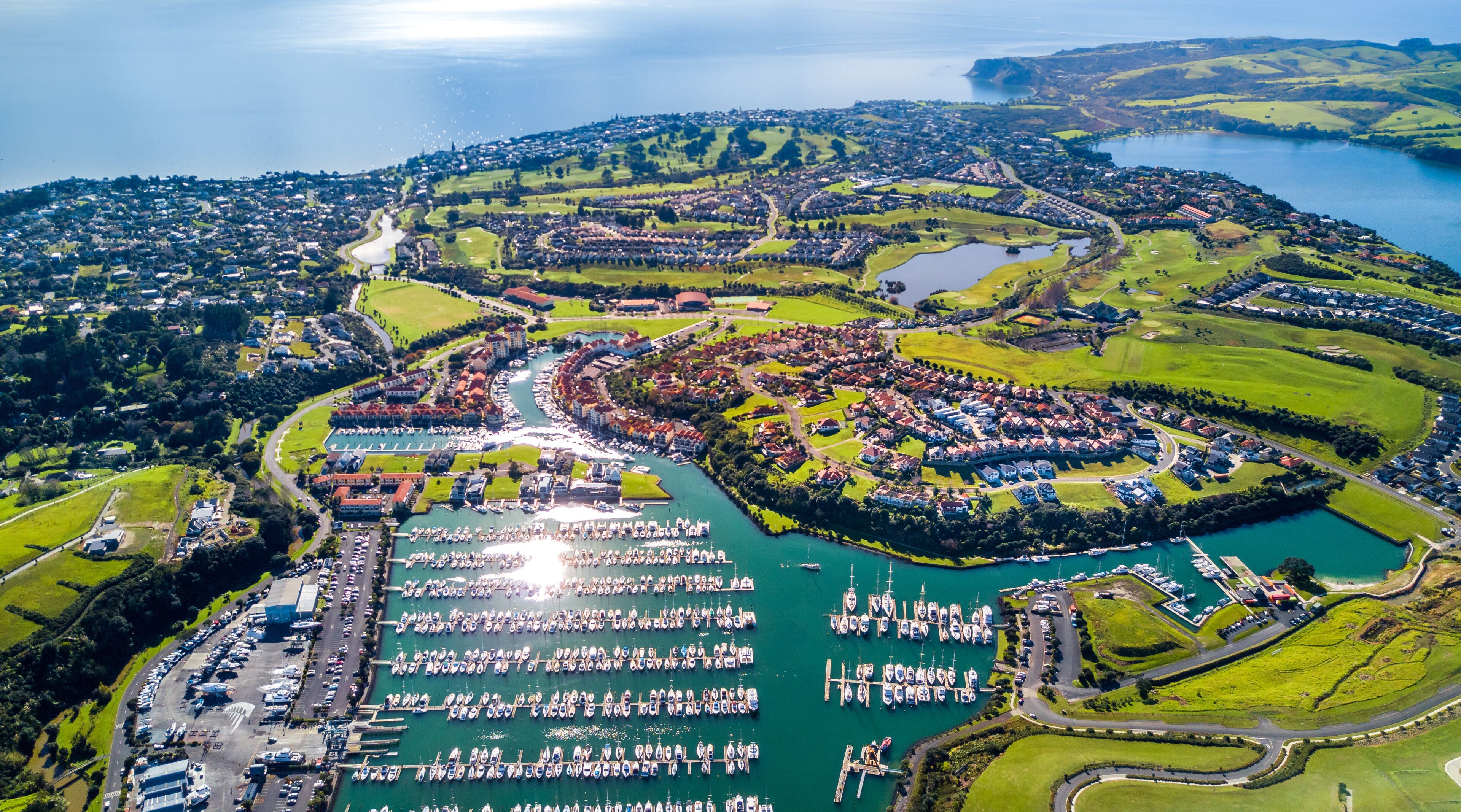 An aerial view of a coastal town with a marina, green hills, and a large body of water.
