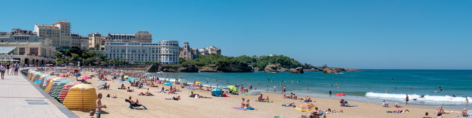 Praia cheia de pessoas se banhando e nadando, com um litoral rochoso e edifícios ao fundo.