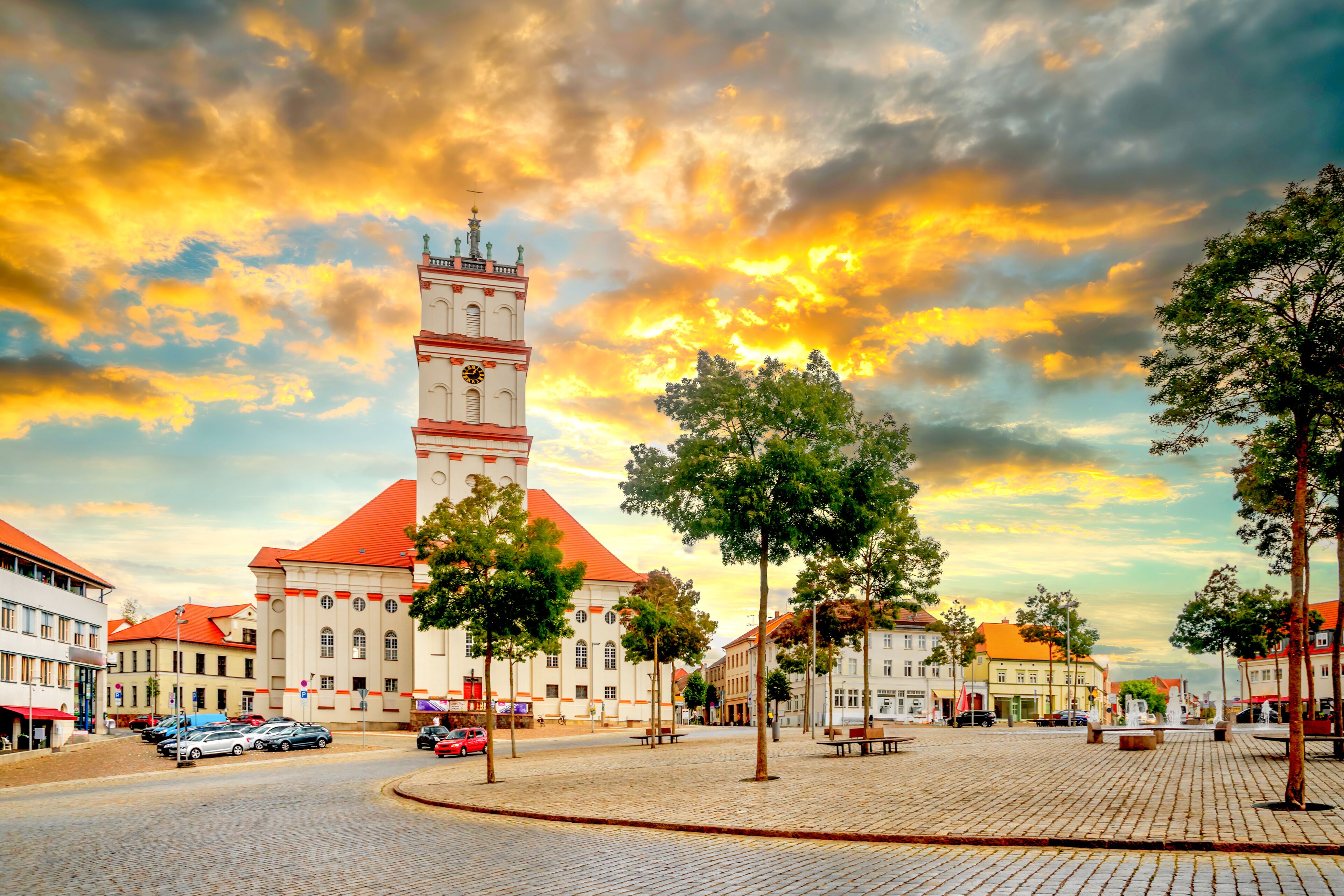 A cobblestone square with a prominent clock tower, surrounded by historic buildings and trees.