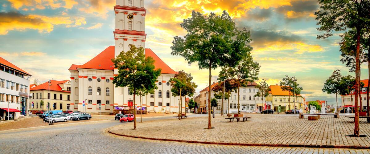 A cobblestone square with a prominent clock tower, surrounded by historic buildings and trees.
