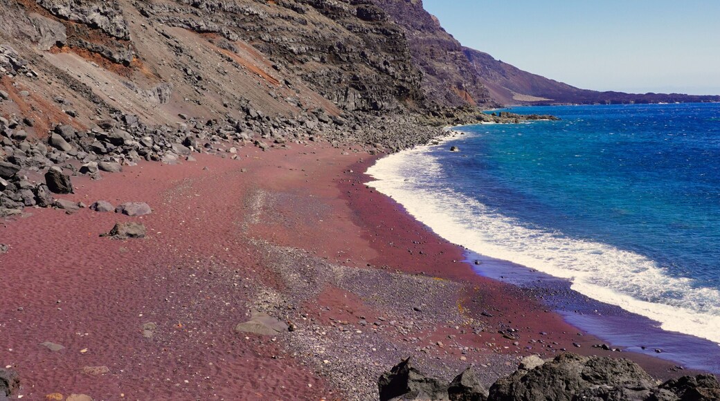 A beach with red sand, a rocky cliff, and clear blue water.