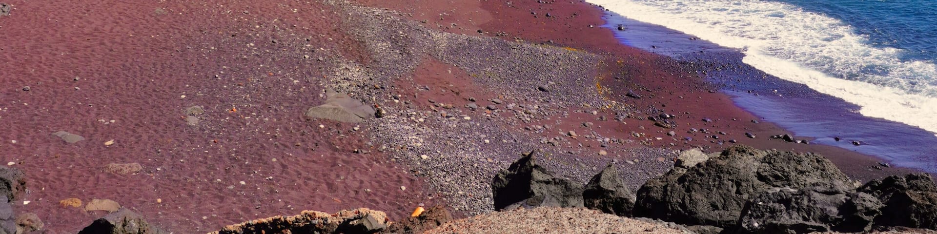 A beach with red sand, a rocky cliff, and clear blue water.