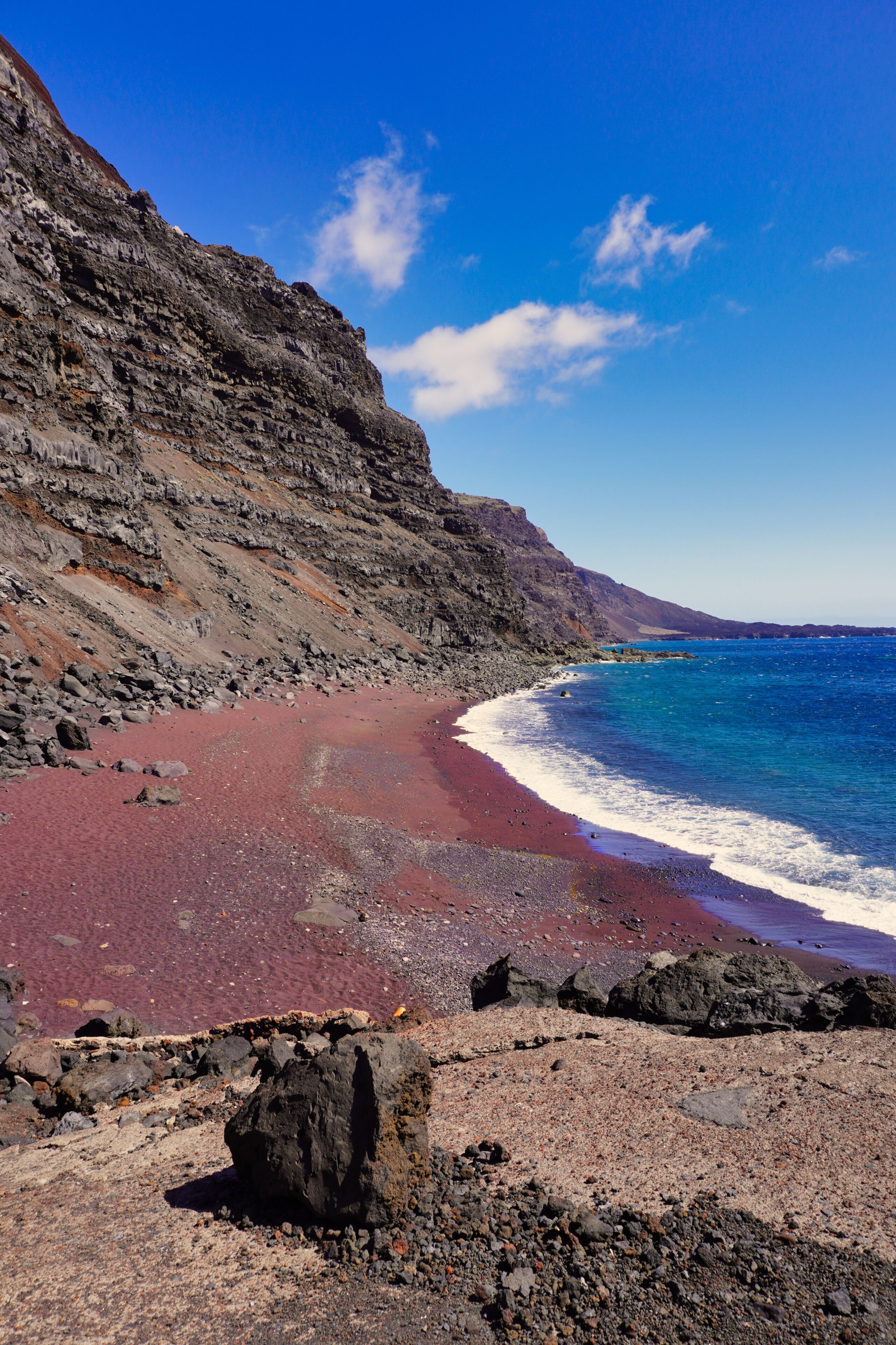 A beach with red sand, a rocky cliff, and clear blue water.