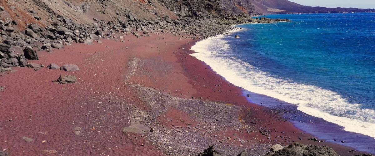 A beach with red sand, a rocky cliff, and clear blue water.