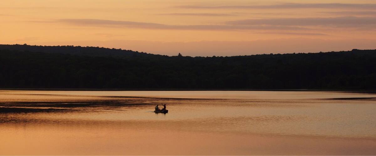 A calm lake at sunset with a silhouette of a boat and a forested hill in the background.
