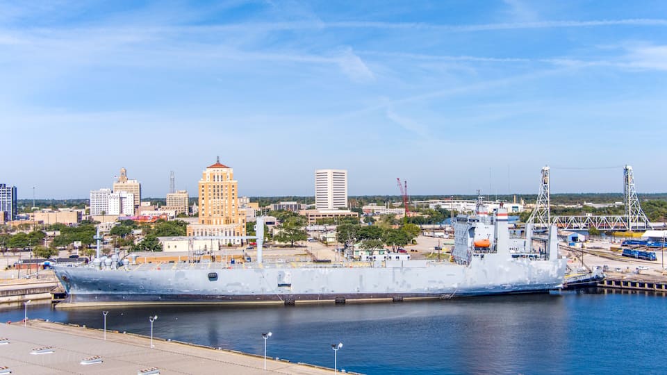 A large naval ship docked at a port with a city skyline in the background.