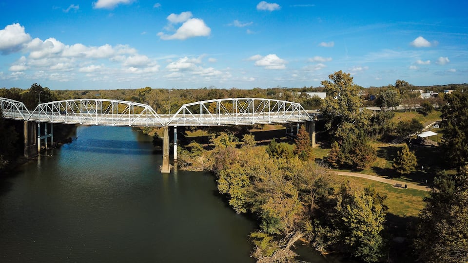 A white metal bridge spanning a river with a park and houses in the background.