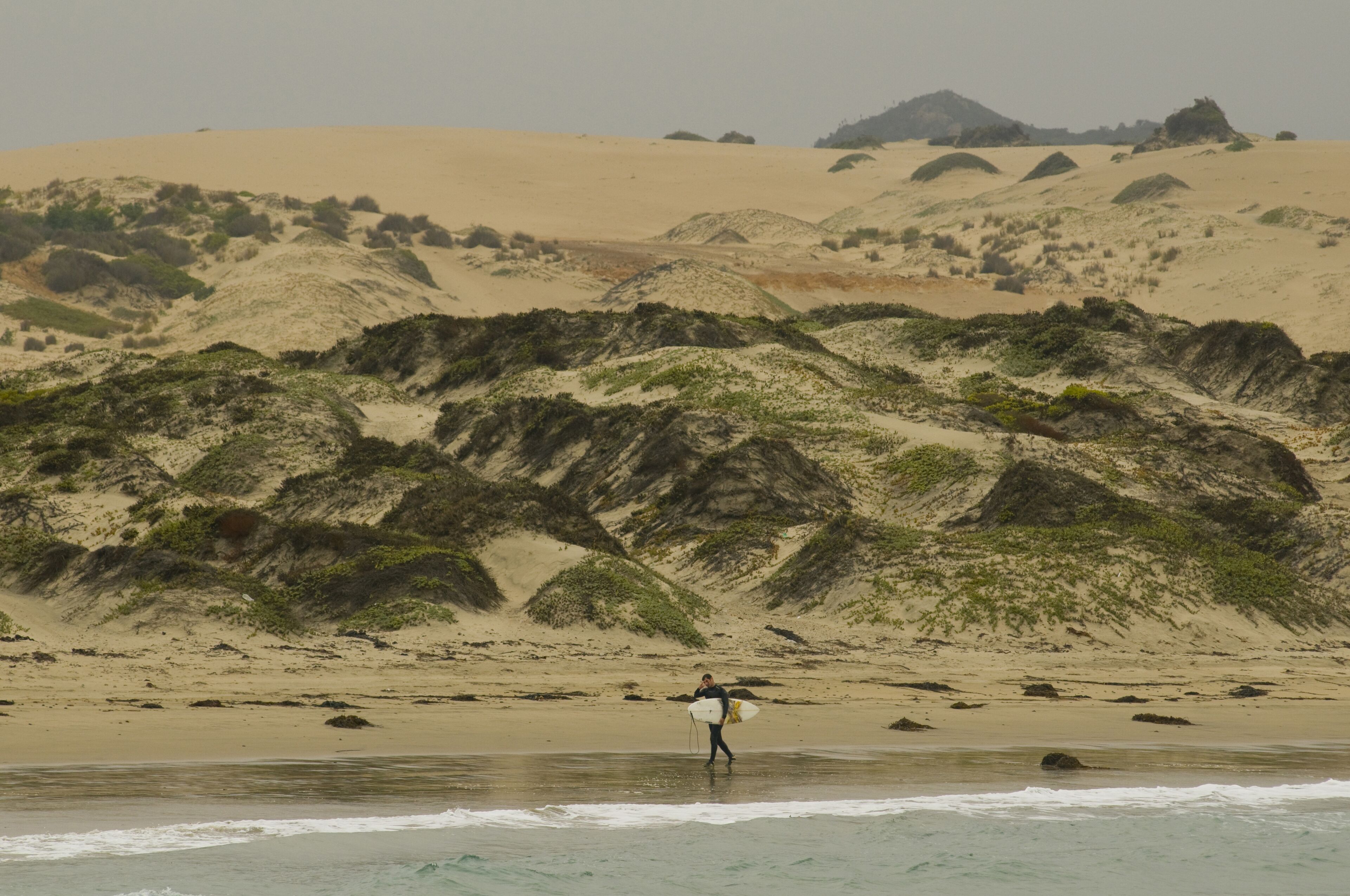 A person with a surfboard on a beach with sand dunes and rocky outcrops.