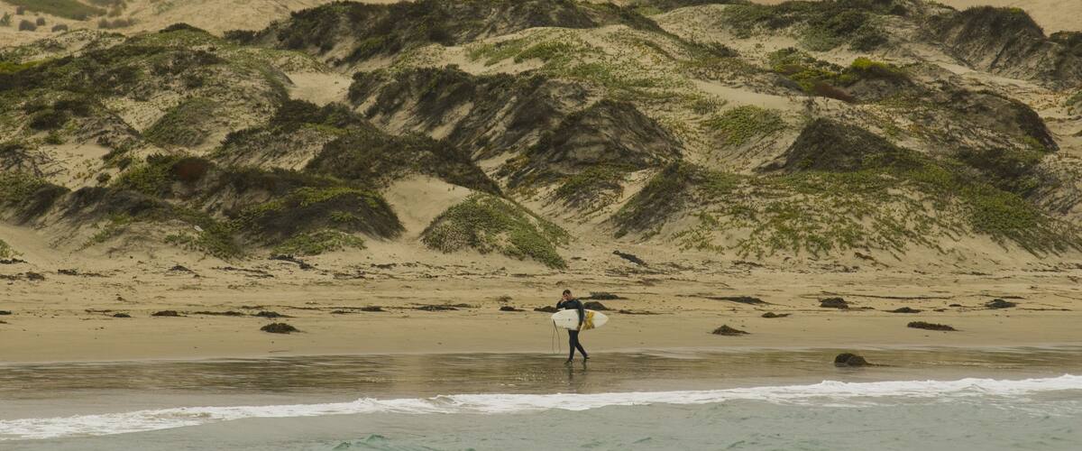 A person with a surfboard on a beach with sand dunes and rocky outcrops.