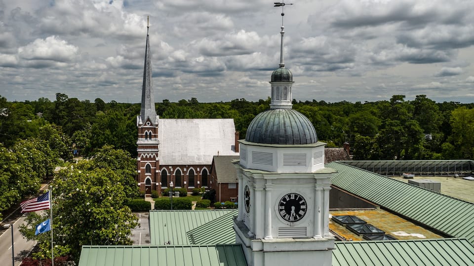 A church with a steeple and a clock tower, surrounded by greenery.