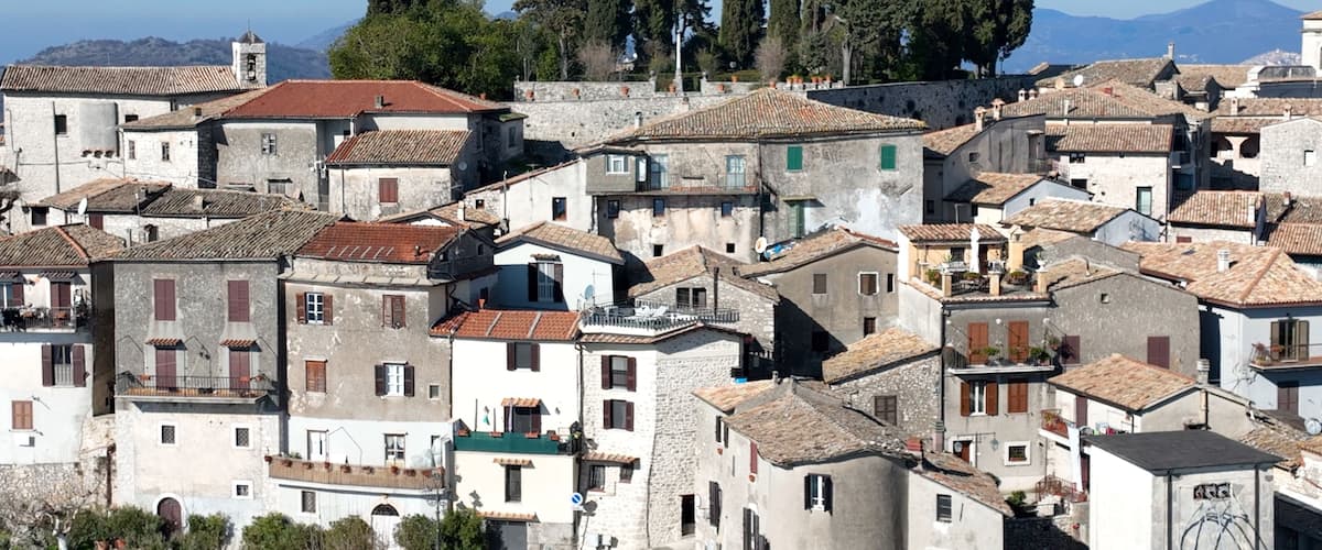 A densely packed historic town with stone buildings, red-tiled roofs, and a backdrop of mountains.