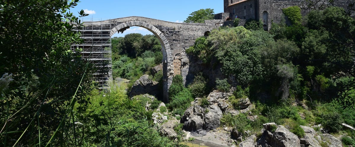 An ancient stone bridge with a tower, surrounded by lush greenery and rocky terrain.