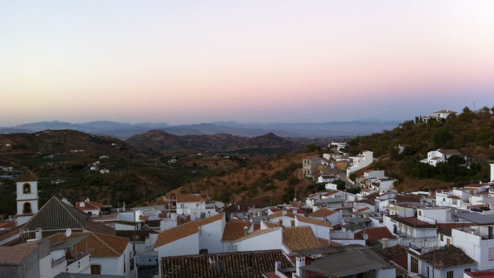 A coastal town with white buildings and terracotta roofs, surrounded by hills and mountains.