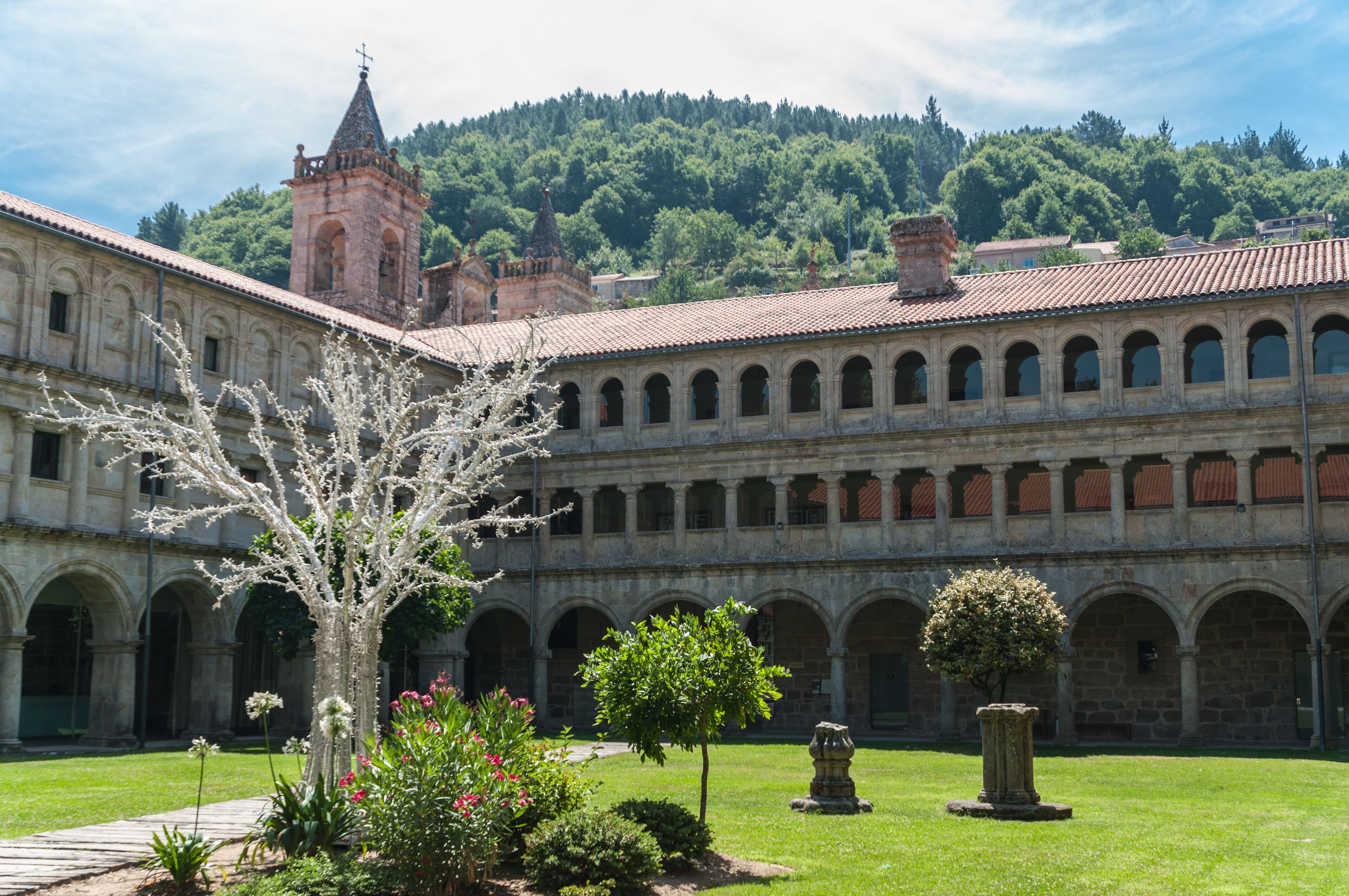 A historic courtyard with a large tree, a stone fountain, and a building with a bell tower.