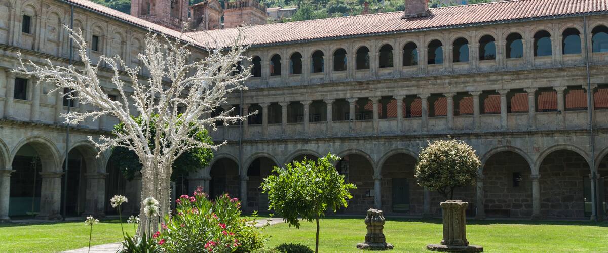 A historic courtyard with a large tree, a stone fountain, and a building with a bell tower.