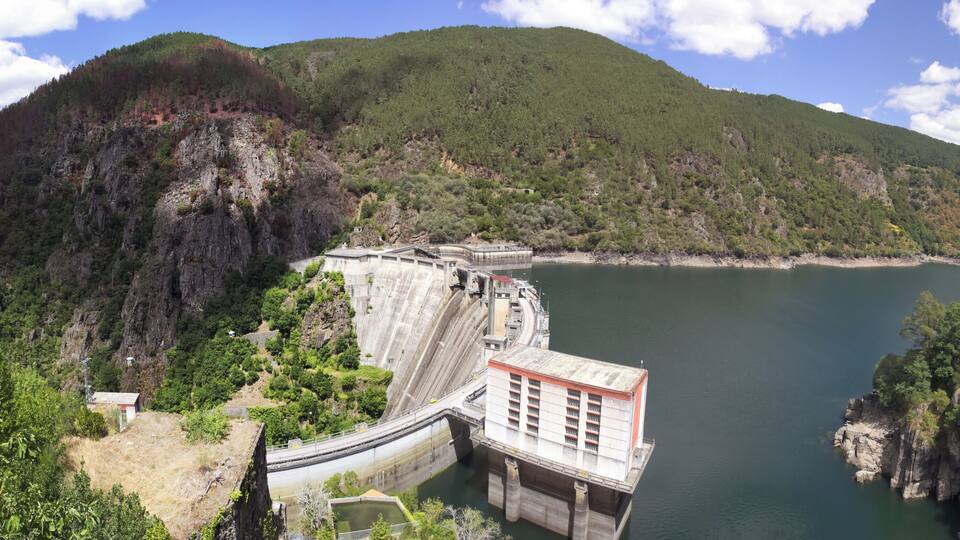 A dam with a lock system, surrounded by forested hills and a calm lake.