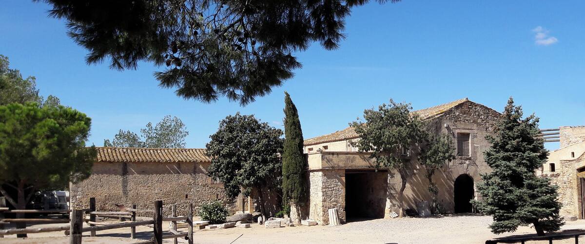 A historic stone building with a tiled roof, surrounded by trees and a gravel courtyard.
