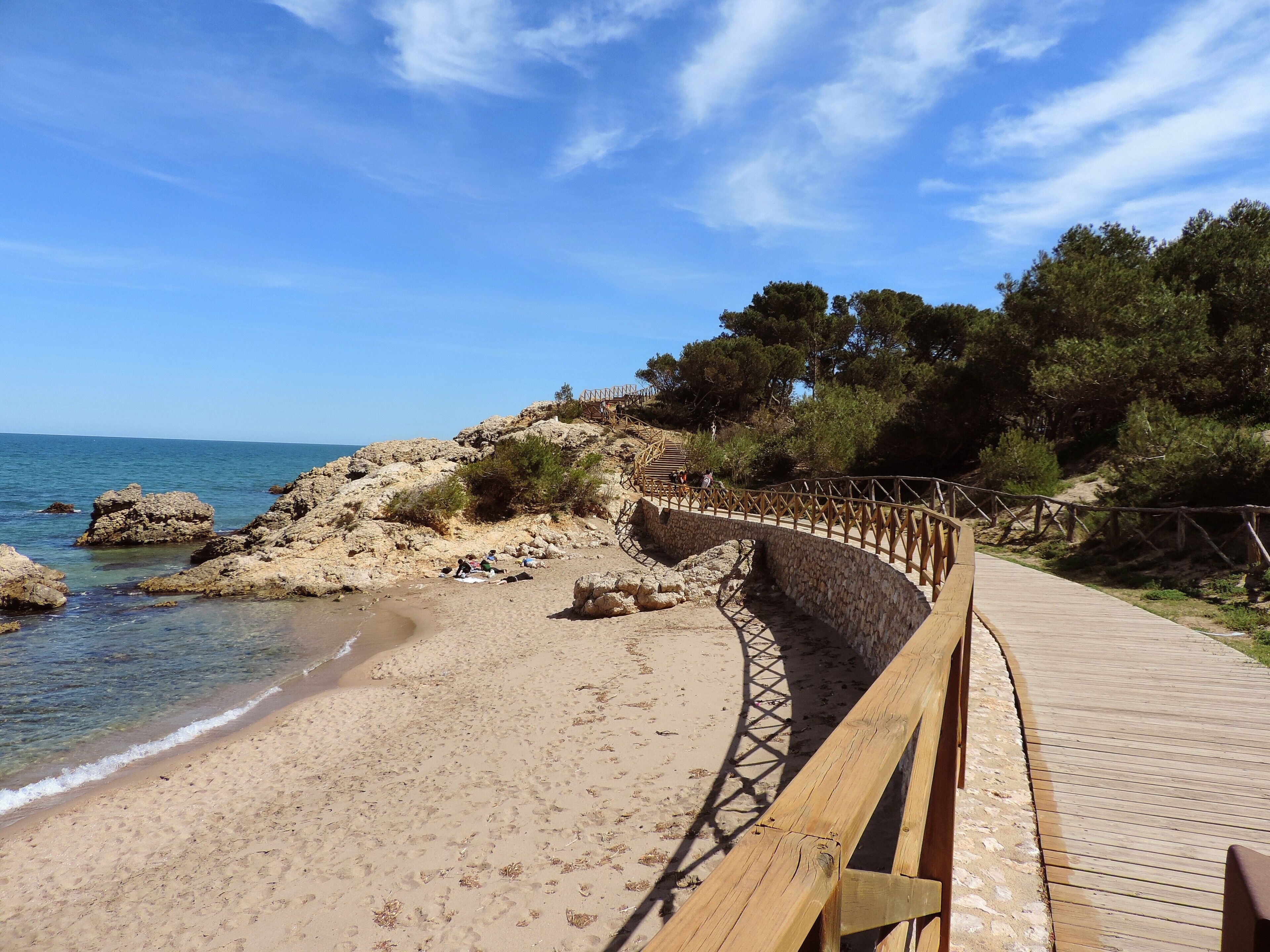 Un paseo marítimo de madera a lo largo de una playa arenosa con afloramientos rocosos y árboles al fondo.
