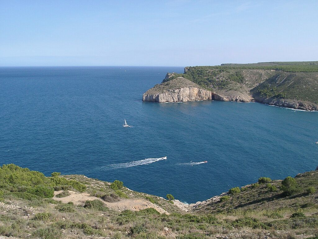 A coastal landscape with cliffs, a clear blue sea, and a sailboat.