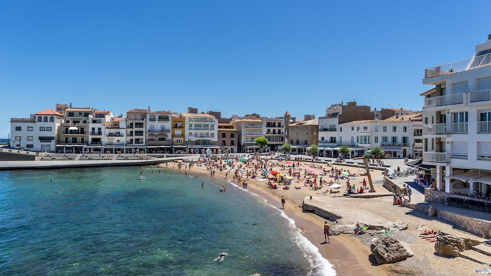 A crowded beach with clear blue water, a rocky shoreline, and buildings along the coast.