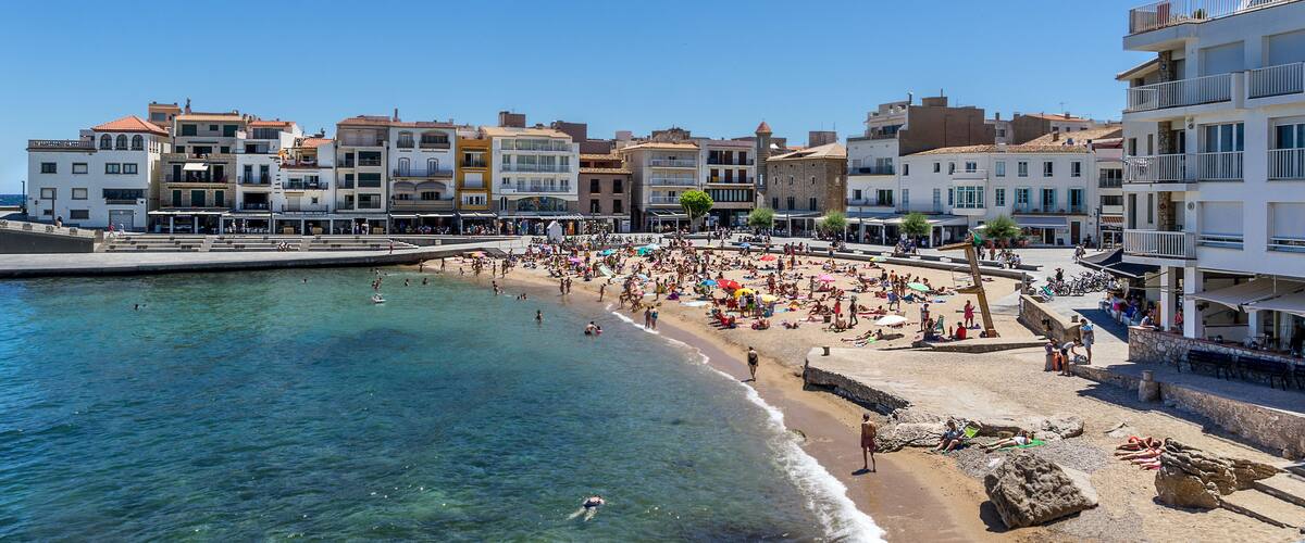 A crowded beach with clear blue water, a rocky shoreline, and buildings along the coast.