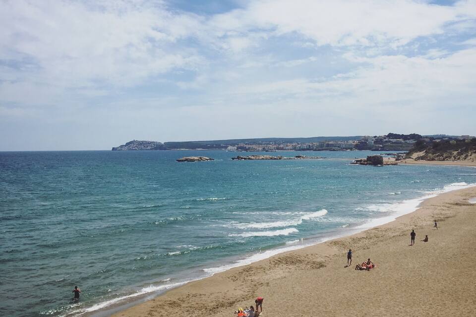 A beach with people sunbathing and swimming, a rocky shoreline, and buildings in the distance.