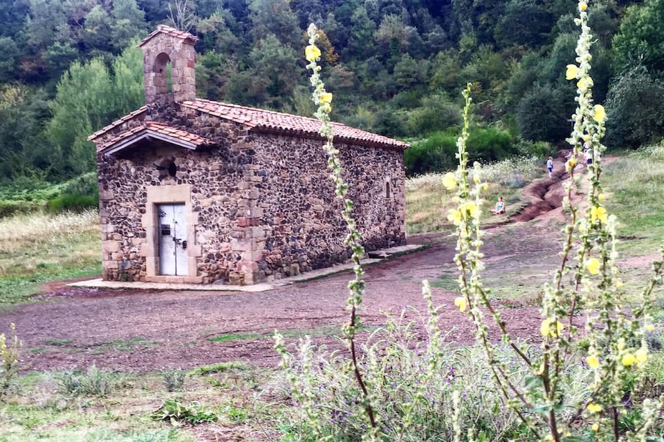 A stone building with a tiled roof, surrounded by a forested hillside.