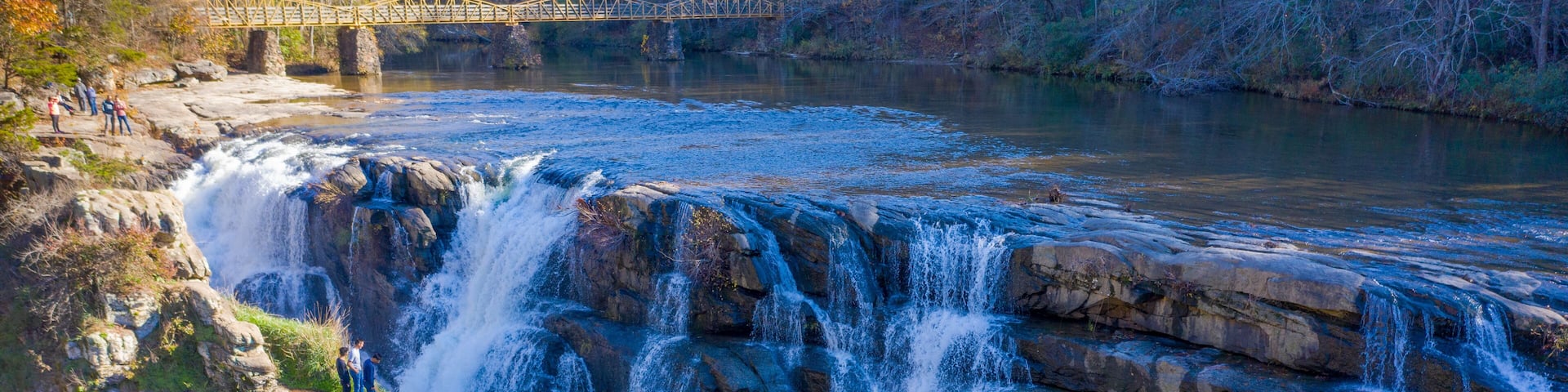 Scenic High Falls Park waterfall in North Alabama with hikers on cliff edge and pedestrian bridge in background
