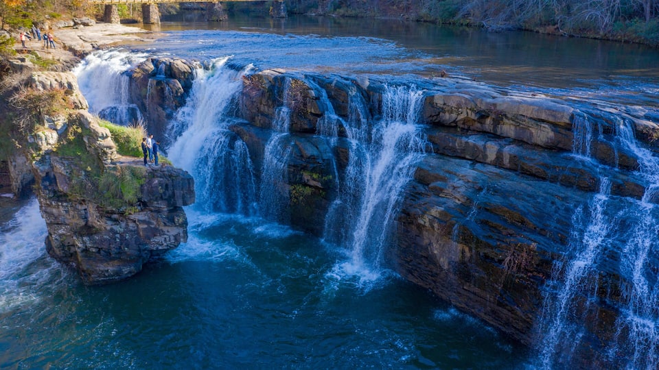 A waterfall cascading over rocks, a bridge in the background, and a group of people observing the scenery.