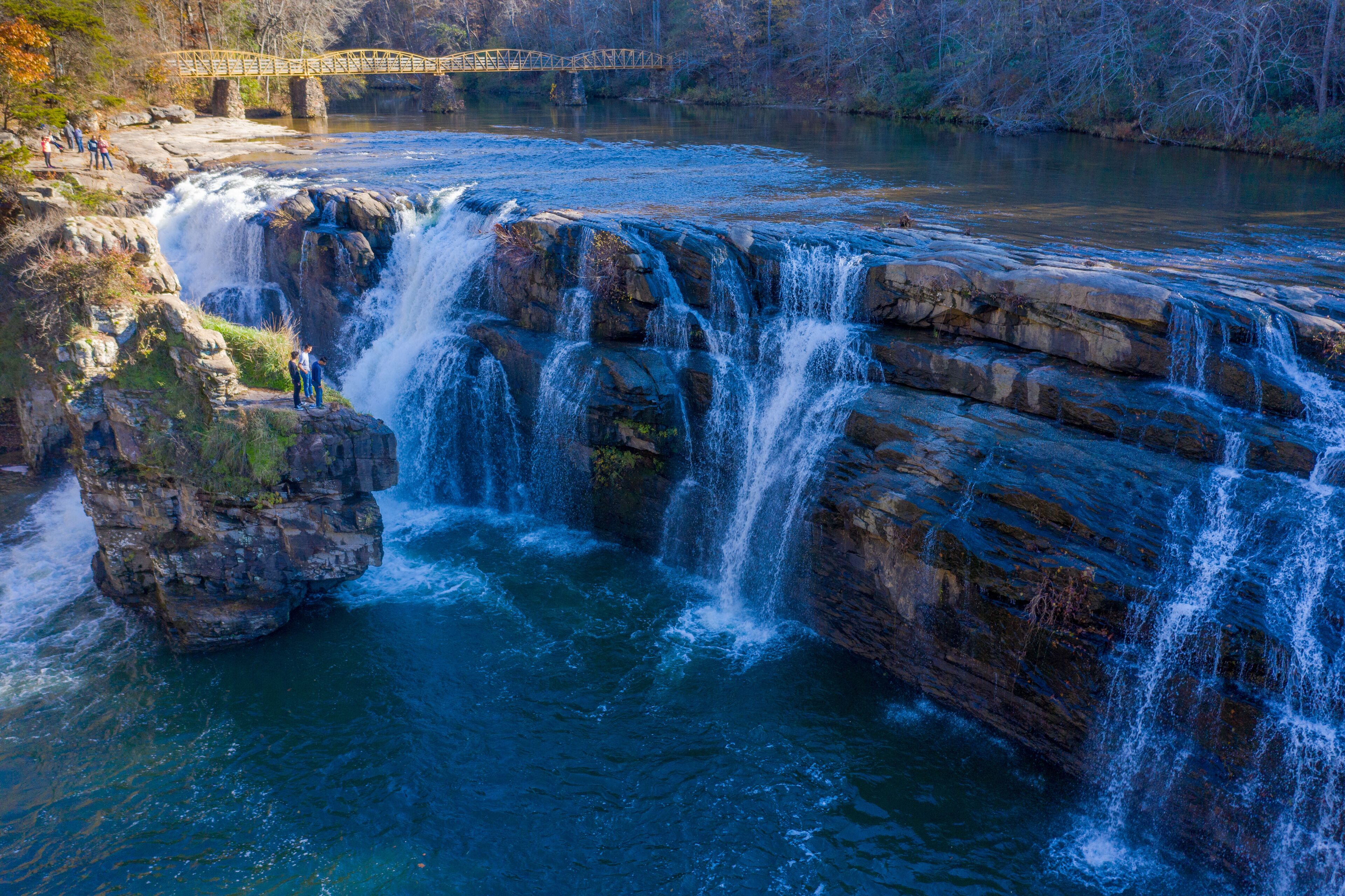 A waterfall cascading over rocks, a bridge in the background, and a group of people observing the scenery.