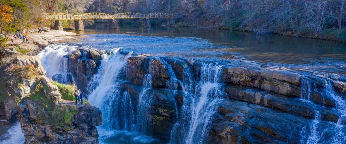 A waterfall cascading over rocks, a bridge in the background, and a group of people observing the scenery.