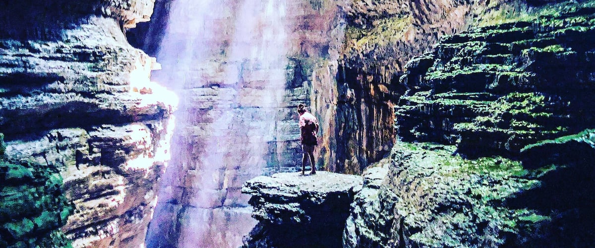 A person standing on a rock formation with a waterfall cascading down a cliff.