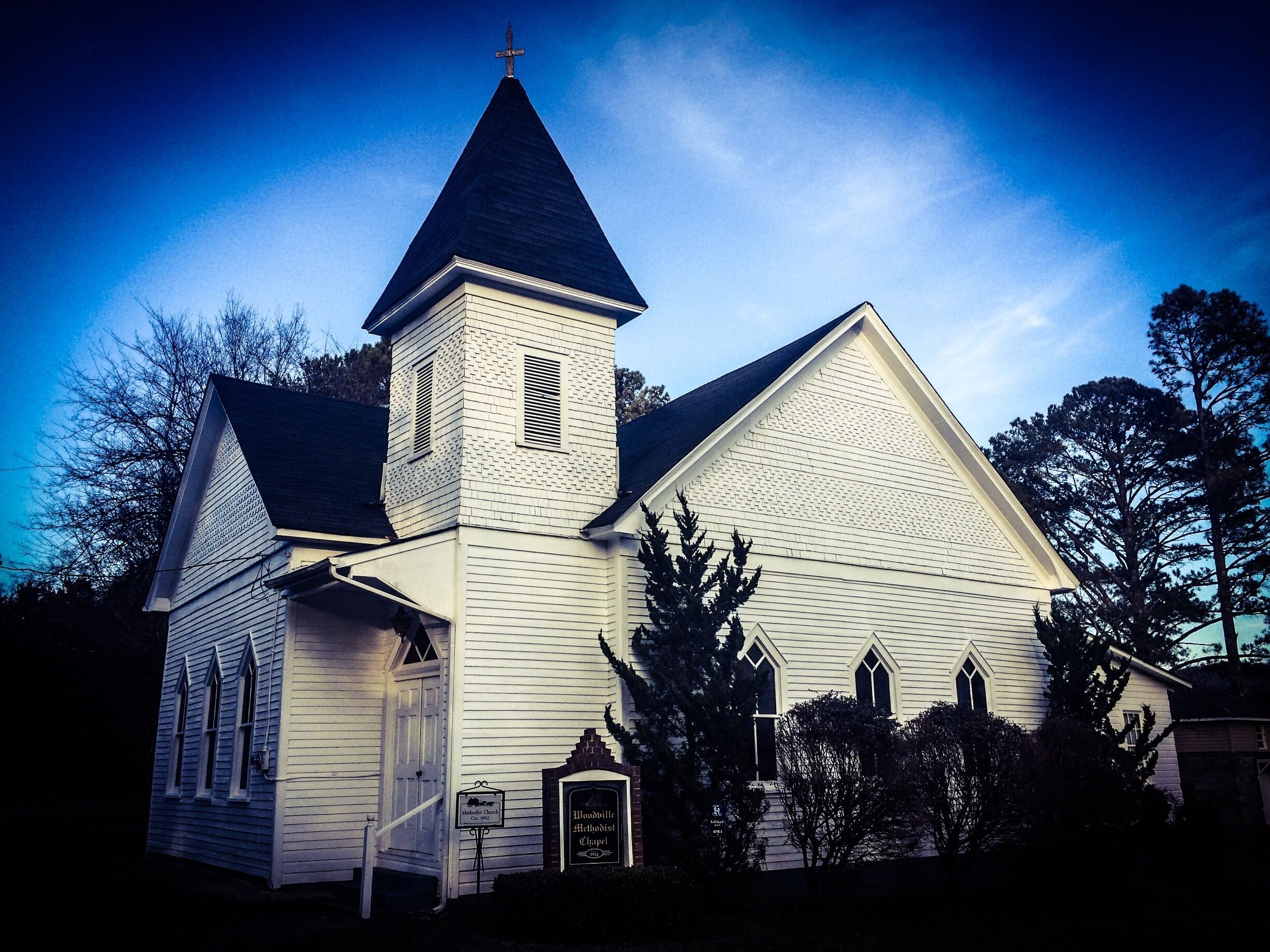 A white church with a steeple and a cross, surrounded by trees.