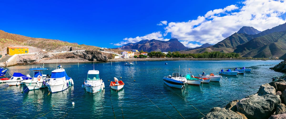 A harbor with boats, rocky shore, and mountains in the background.